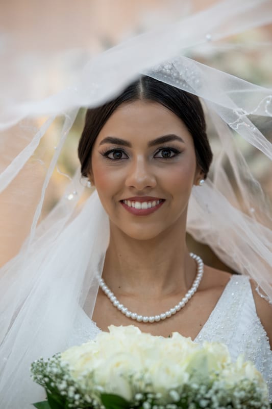 Noiva sorrindo sob o véu, com colar de pérolas e buquê branco